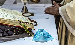 Ein Priester faltet die Hände und feiert den Gottesdienst am Altar, auf dem das Messbuch aufgeschlagen liegt, daneben eine einfache Atemschutzmaske, dahinter liturgische Geräte, am 8. Mai 2020 in Rom.