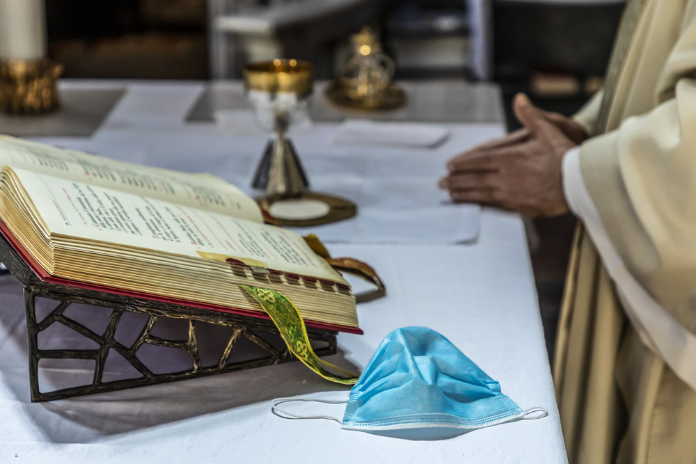 Ein Priester faltet die Hände und feiert den Gottesdienst am Altar, auf dem das Messbuch aufgeschlagen liegt, daneben eine einfache Atemschutzmaske, dahinter liturgische Geräte, am 8. Mai 2020 in Rom.