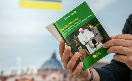 Eine Person hält das Nachsynodale Apostolische Schreiben 'Querida Amazonia' von Papst Franziskus zur Amazonas-Synode in der Sprache Italienisch am 12. Februar 2020 auf einem Tisch im Vatikan. Im Hintergrund: ein Bild vom Petersdom.
